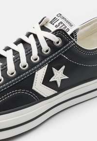 Svart lädersneaker med vit gummikydd. Har vita snören, sömmar och en stjärnlogotyp i en kontrasterande färg. Klassisk design.