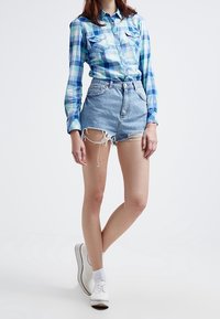 Camicia a quadri blu chiaro con bottoni e due taschini sul petto, abbinata a pantaloni corti di jeans a vita alta con orlo sfibrato. Le scarpe e le calze bianche completano il look.