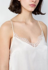 Camisola in seta color crema con spalline sottili e scollatura in pizzo ondulato. Il tessuto ha una texture liscia e una leggera lucentezza.