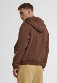 Brauner Hoodie mit lässiger Passform, ausgestattet mit einer Kapuze mit Kordelzug, gerippten Bündchen und Saum. Glatte Textur, kombiniert mit hellbeigen Hosen.
