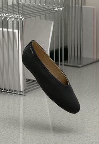 Zwarte Calvin Klein platte schoen met een ronde neus zwevend nabij een metalen file-organizer op een grijspikkelachtige ondergrond.