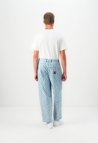 Man standing with back facing viewer, wearing white t-shirt, light blue polka dot jeans, and white sneakers against a plain white background.