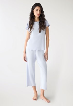 Donna con lunghi capelli scuri e mossi, che indossa un top azzurro chiaro a maniche corte e pantaloni larghi corti, in piedi scalza su uno sfondo bianco semplice.