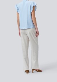 Mujer de pie con una blusa corta de mangas con volantes de color azul claro, pantalones rectos en rayas verticales beige y blancos, y sandalias de tacón marrón, de espaldas.