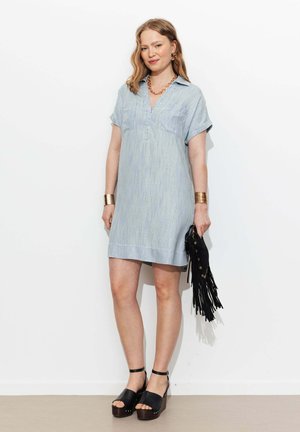 Femme portant une robe bleu clair à manches courtes et des sandales compensées noires, tenant un sac à main noir à franges, debout contre un mur blanc.