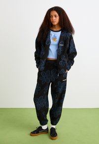 Giacca e pantaloni in pile neri con motivi floreali blu, abbinati a un crop top blu chiaro con un design ricamato e sneaker neri.