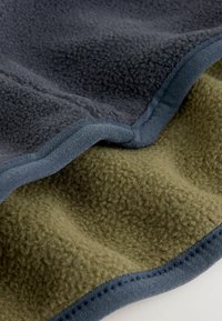 Fleece tæppe med et mørkegråt toplag og en olivengrøn underside. Har en blød tekstur og kontrasterende blå kanter langs sømmene.