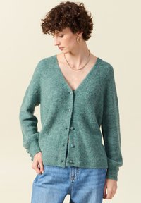 Grön stickad cardigan med V-ringning, knäppning fram och långa ärmar. Mjuk textur och avslappnad passform, ihop med blå jeans.