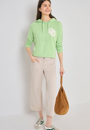 Mujer con sudadera con capucha verde claro y pantalones beige anchos, sosteniendo un bolso marrón de ante en su mano derecha, de pie contra una pared blanca.