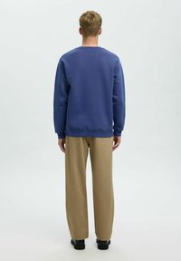 Mėlynas džemperis ilgomis rankovėmis ir pūstomis apačiomis derinamas su smėlio spalvos plačiomis kelnėmis. Modelis matomas iš nugaros.