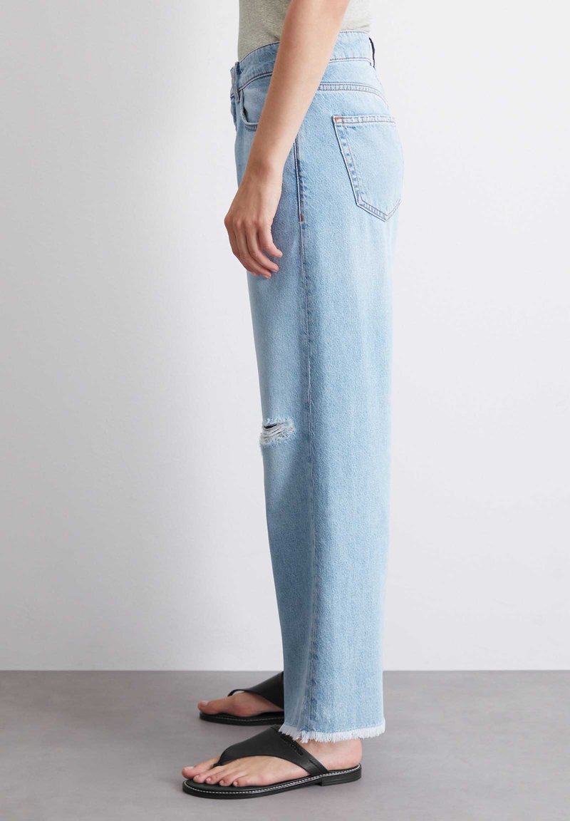 Jasnoniebieskie dżinsy o prostych nogawkach, z przetarciami, postrzępionym dołem i średnim stanem. Noszone z czarnymi klapkami.