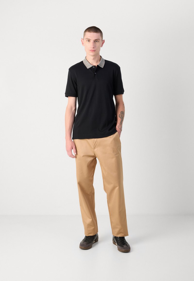Schwarzes Poloshirt mit einem gemusterten Kragen, kombiniert mit beigen Chinos und dunklen Schuhen. Minimalistisches Design und figurbetonte Silhouette.
