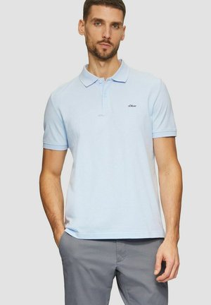 Mann trägt ein hellblaues Poloshirt mit kurzen Ärmeln und graue Hose, steht mit einer Hand in der Tasche vor einem schlichten Hintergrund.