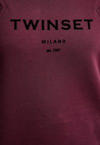 Bordový pulóver s rebrovaným výstrihom a vyšitým nápisom "TWINESET MILANO est. 1987" v čiernej farbe, s hladkou, textúrovanou látkou.