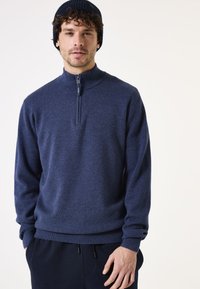 Marineblauwe half-zip trui met een gestructureerde brei en geribbelde mouwen. Draag met donkere broeken, handen in de zakken.