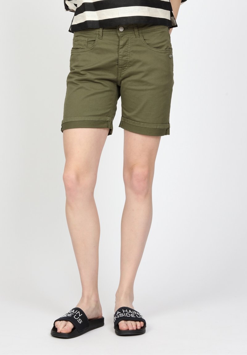 Olivgröna uppvikta shorts med framfickor, metallknäppning och kontrasterande svarta sandaler med vit text.