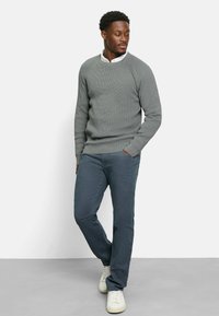 Grau gestrickter Pullover mit rundem Halsausschnitt, kombiniert mit dunkelblauen Hosen und weißen Sneakers. Der Pullover hat eine strukturierte Oberfläche und gerippte Bündchen.