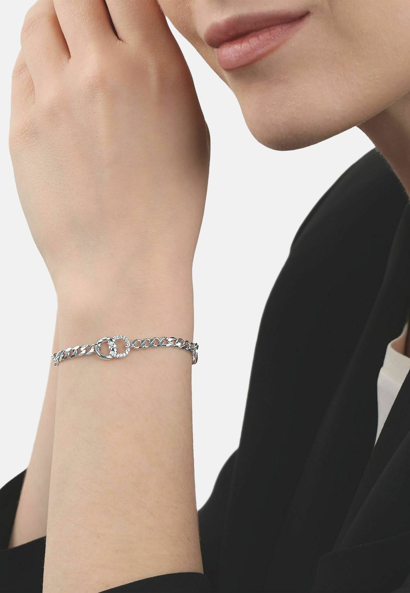 Femme portant un bracelet en chaîne argentée avec deux anneaux entrelacés, dont un incrusté de petits cristaux, à son poignet.