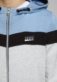 Felpa con zip grigia caratterizzata da blocchi di colore triangolari in azzurro chiaro, nero e grigio. Include una toppa con logo, un cappuccio regolabile con coulisse e un tessuto testurizzato.