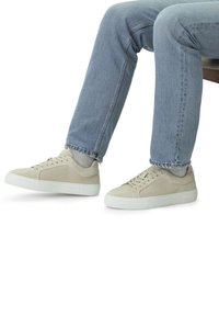 Beige suede sneakers with a low profile and white rubber soles. Laces and side detailing in matching beige. Paired with light blue jeans.