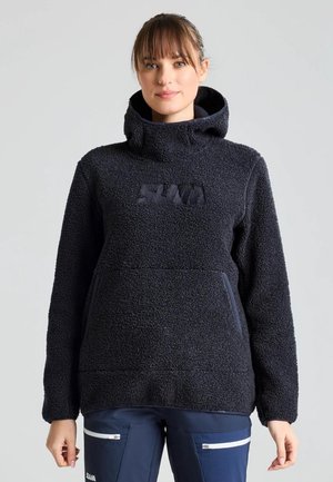 Una mujer luce un jersey polar oscuro con cuello alto y bolsillo delantero, combinado con pantalones azul marino sobre un fondo claro y liso.