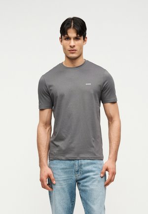 Jeune homme portant un T-shirt à manches courtes gris foncé uni avec le texte « HUGO » et un jean bleu clair, debout devant un fond neutre.