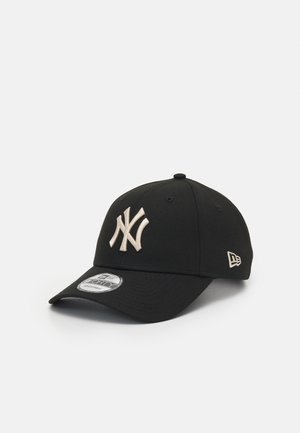 Sort baseballkasket med hvidt New York Yankees-logo foran og lille hvidt mærkelogo på siden, hvilende på en hvid overflade.