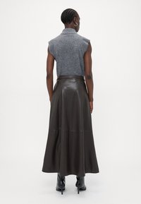 Maglione grigio senza maniche abbinato a una lunga gonna di pelle marrone, con una texture liscia e design svasato. Stivaletti neri alla caviglia completano il look.