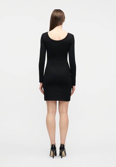 Γυναίκα που στέκεται γυρισμένη πίσω, φοράει εφαρμοστό μαύρο φόρεμα με μακριά μανίκια και μαύρα παπούτσια με ψηλό τακούνι σε απλό λευκό φόντο.