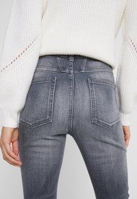 Jeans de denim gris ajustados con una cintura alta, caracterizados por dos bolsillos en la parte trasera y una textura suave, combinados con un suéter de punto blanco.