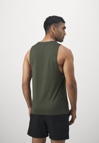 Tirante en tela verde oliva con una textura sutil, que presenta un pequeño logo negro en la parte trasera, combinado con pantalones cortos negros.