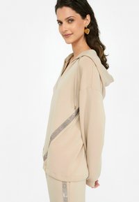 Sweatshirt beige à capuche avec un décolleté en V, panneau latéral avec un accent métallique et des manches amples. Texture de tissu lisse.