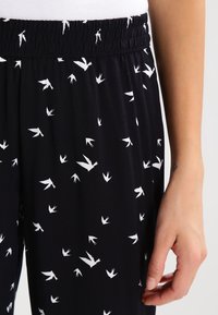 Černé látkové kalhoty se vzorem bílých ptáků. Elastický pas; hladký, lehký materiál. Detailní záběr ruky držící kalhoty.