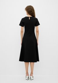Schwarzes knie-langes Kleid mit kurzen Ärmeln, A-Linie, Rückenschlitz und glatter Textur. Getragen mit silbernen High Heels.