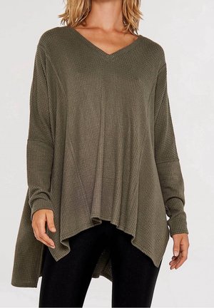 Femme portant un pull en maille gaufrée vert olive ample avec un col en V et un ourlet asymétrique, associé à un pantalon noir.
