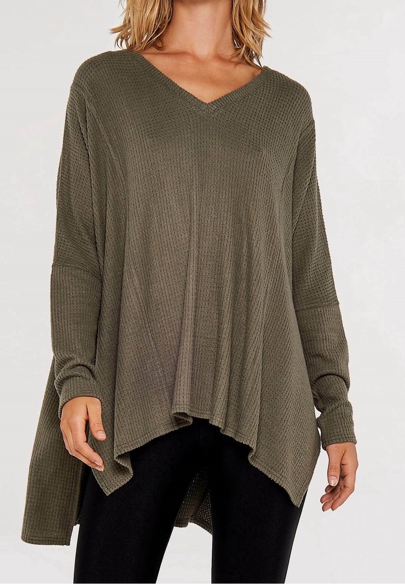 Femme portant un pull en maille gaufrée vert olive ample avec un col en V et un ourlet asymétrique, associé à un pantalon noir.