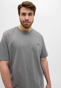 Camiseta de algodón gris con mangas cortas y cuello redondo. Presenta un pequeño logo en el lado izquierdo. Textura suave, ajuste relajado.
