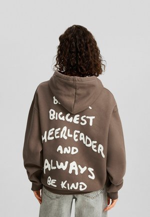 Personne aux cheveux bouclés portant un sweat à capuche marron avec le texte "Biggest Cheerleader and Always Be Kind" dans le dos, debout devant un fond uni.