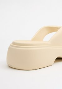 Sandalia de plataforma beige con una tira ancha y un acabado texturizado. Suela redondeada y ligera elevación en el tacón. Material suave y mate.