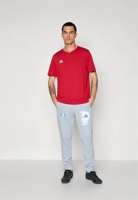 Man staat met handen in zijn zakken, draagt een rood Adidas T-shirt, lichtgrijze broek met grafische logo's en zwarte sneakers tegen een effen achtergrond.