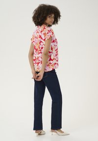 Kratkoročnovrata cvetna bluza v roza in oranžni barvi, v kombinaciji s temno modrimi jeansi. Model stoji z rokami sklenjenimi za hrbtom, obut v bež pete.