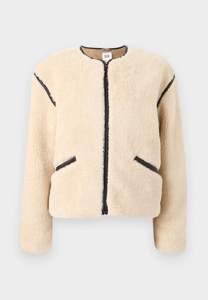 Veste en fausse fourrure beige avec un col rond, une fermeture éclair à l'avant, des accents noirs sur les côtés, et deux poches latérales. Matière douce et texturée.