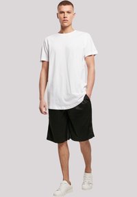 White cotton t-shirt with short sleeves paired with black athletic shorts. The model wears white sneakers, standing against a neutral background.