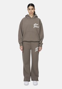 Bruine hoodie en bijpassende sweatpants gemaakt van zachte stof. Bevat een voorkantzak en witte tekstdetails op beide stukken.