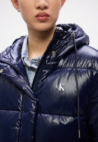 Jachetă puffer albastră cu un finisaj lucios, design umplut, închidere cu fermoar, glugă și accent cu logo. Are o cămașă din denim dedesubt.