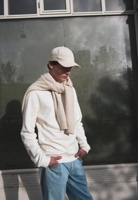 Felpa bianca, sciarpa beige chiaro posata sulle spalle, jeans azzurri chiari e cappellino beige chiaro. Texture morbide, design casual.