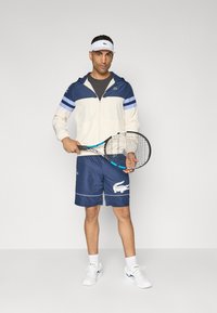 Férfi sportöltözet, amely krém és sötétkék cipzáros dzsekiből, sötétkék rövidnadrágból fehér krokodil logóval és fehér sportcipőből áll. Teniszütőt tart a kezében.