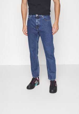Man draagt blauwe recht gesneden spijkerbroek, zwarte sneakers met roze en turquoise accenten en een zwart T-shirt tegen een effen witte achtergrond.