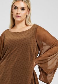 Blusa trasparente marrone con scollatura rotonda e maniche lunghe e fluide. Il tessuto presenta piccoli motivi a pois neri.