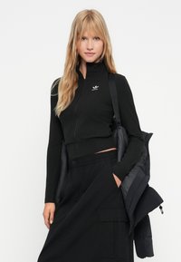 Mladá žena s dlouhými blond vlasy, oblečená v černém zipovém topu Adidas, černých cargo kalhotách a s černou péřovou bundou přes rameno.